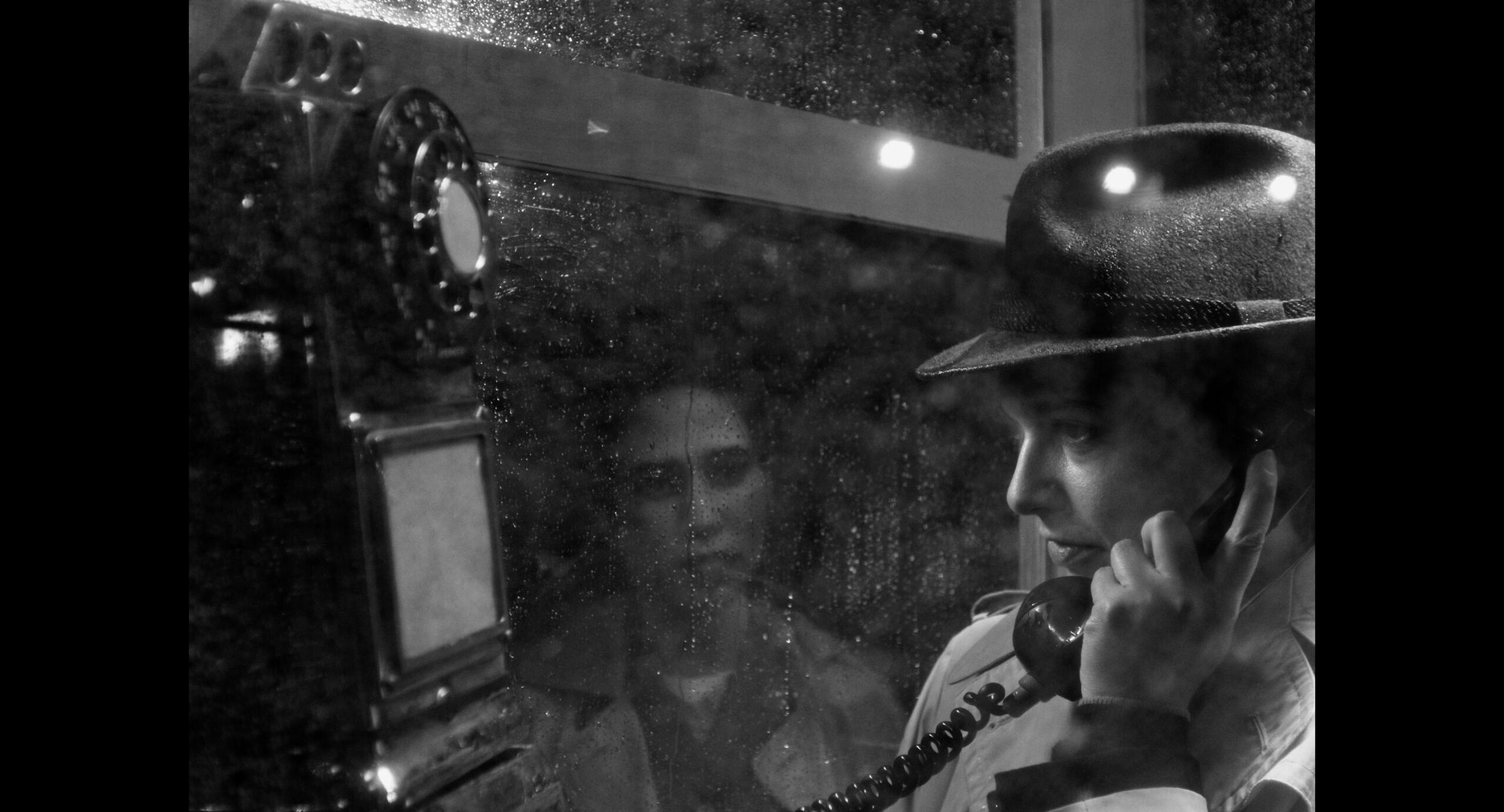 Untitled Butch talks nervously in a phone booth while a zombie-like woman stares at them while standing in the rain.